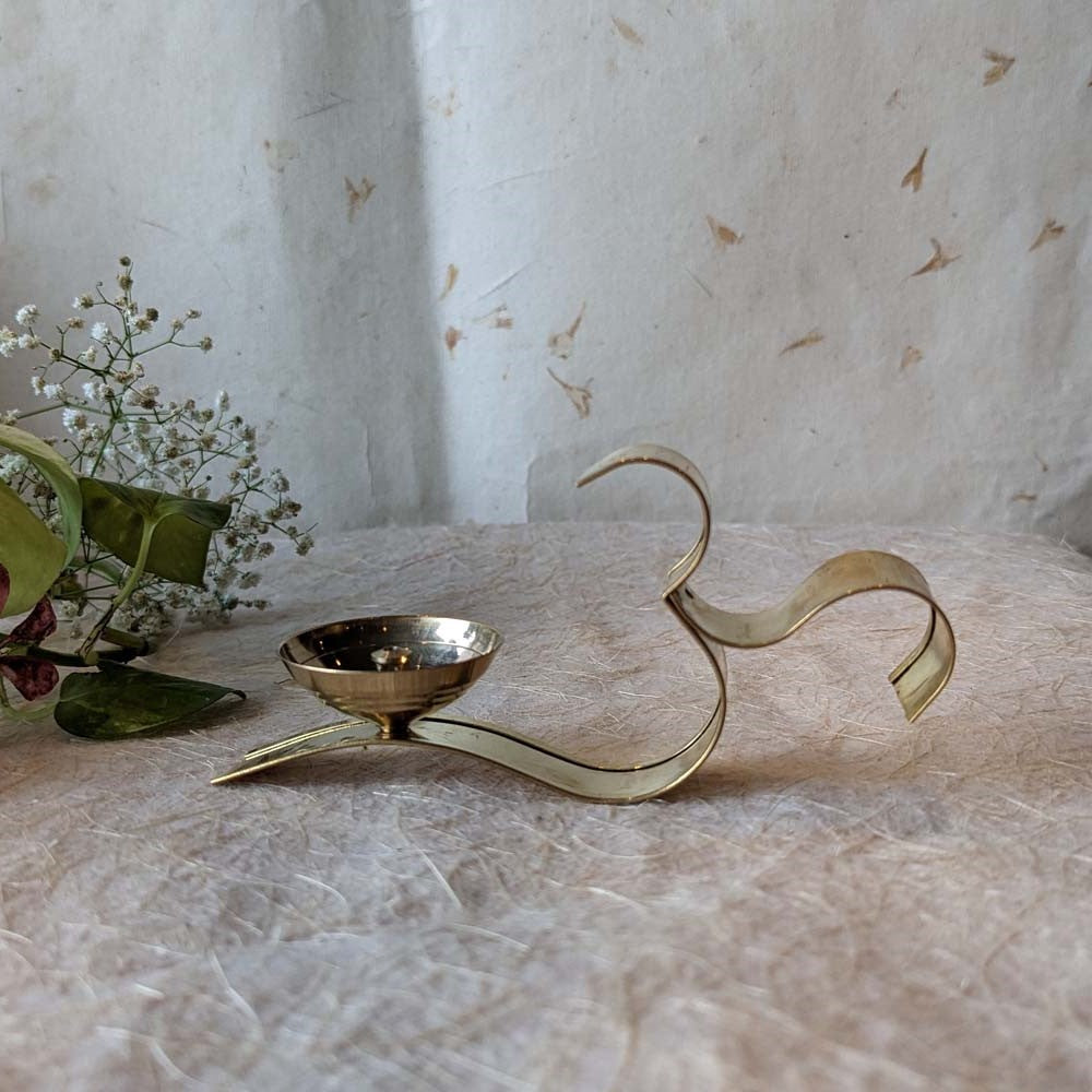Handcrafted brass Om-shaped diya with a polished finish, placed on a textured surface with floral accents.