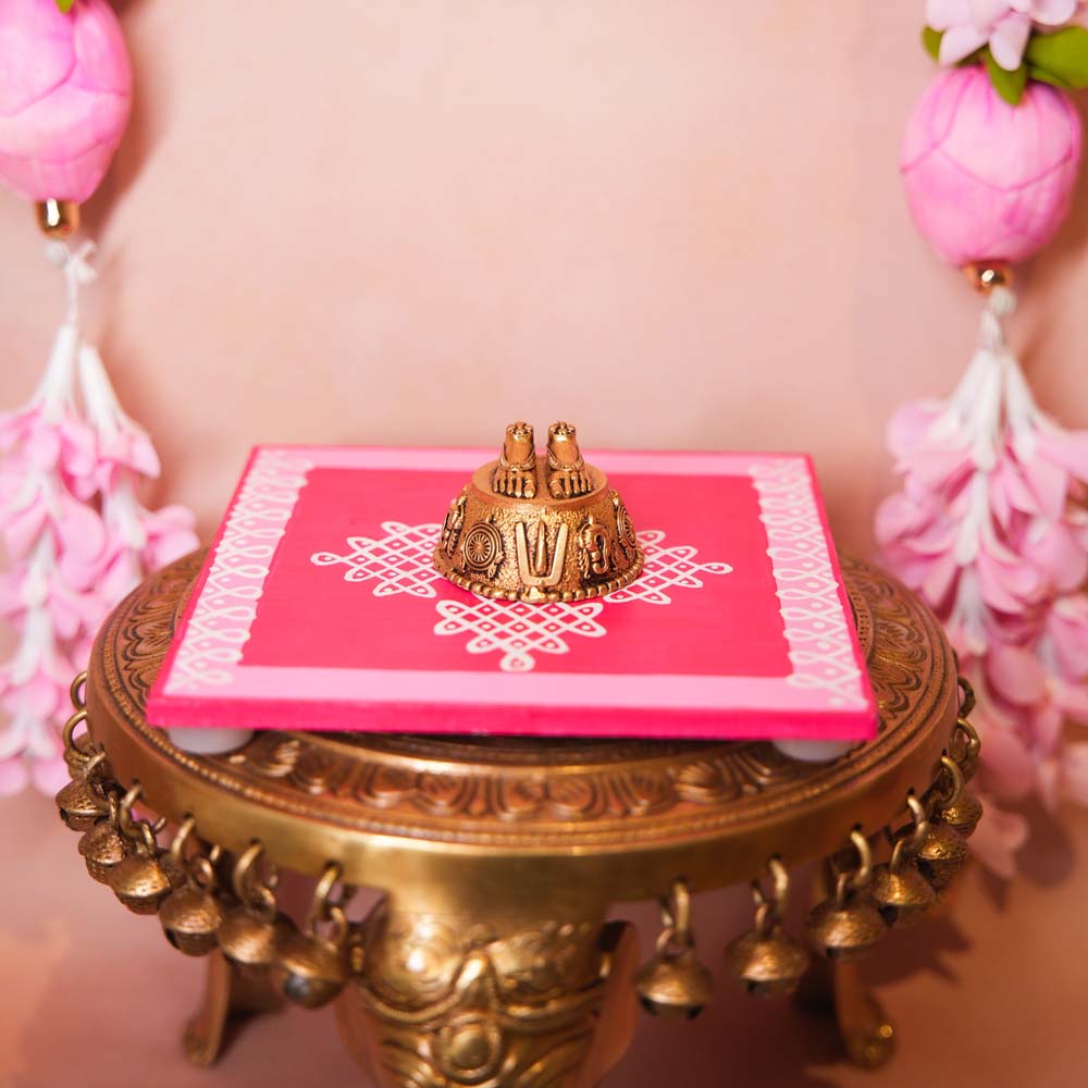 Brass Paduka (Perumal Padam) representing Lord Vishnu's feet, placed on a pink decorative base with traditional kolam design.