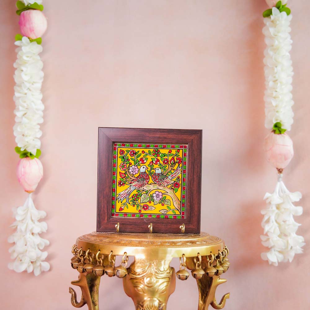 Decorative keyholder featuring a hand-painted motif of a pair of birds on a vibrant yellow background, framed with dark wood, and equipped with key hooks.