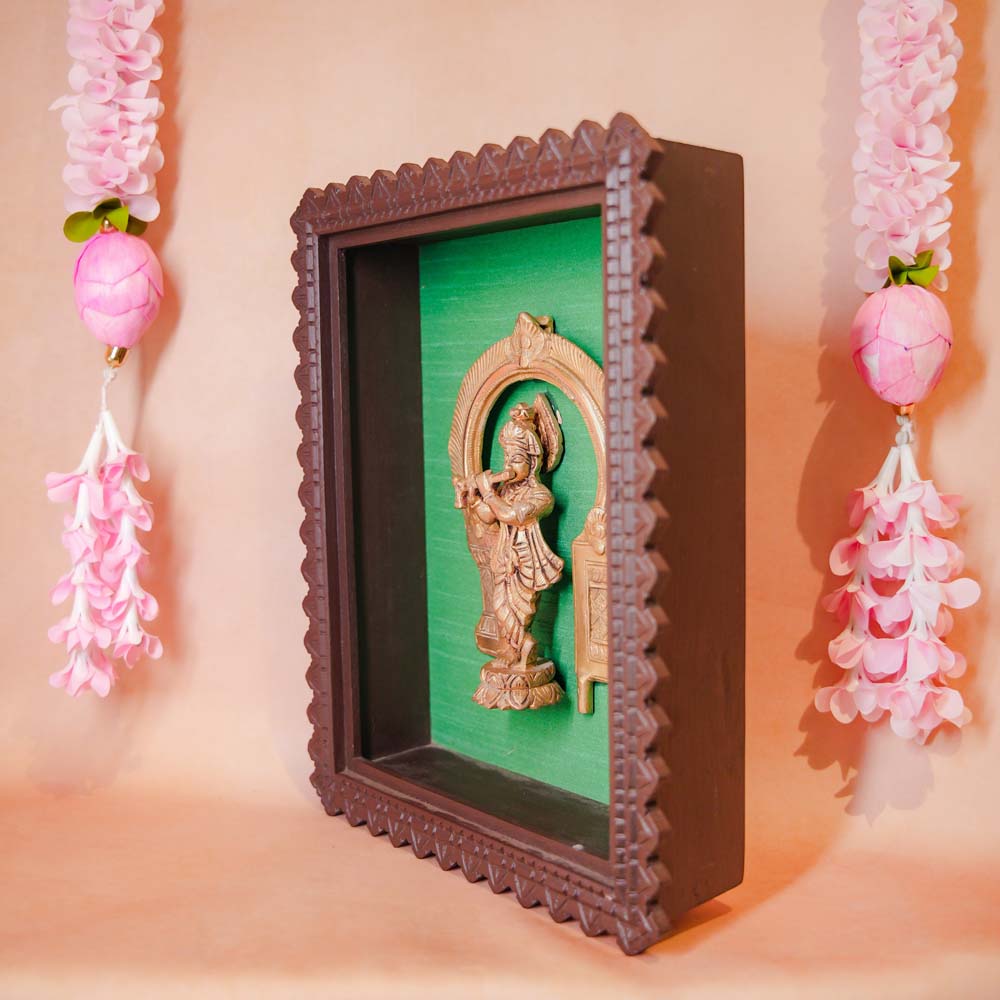 Framed brass sculpture of Lord Krishna playing the flute under an ornate arch, mounted on a green background with a carved wooden frame.