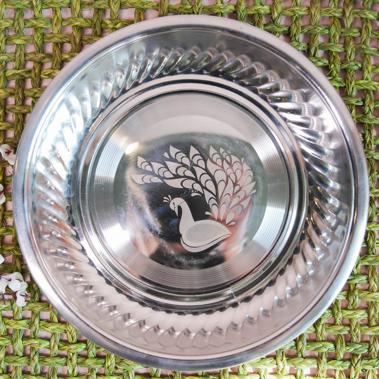 Stainless steel plate with an etched peacock design in the center and a contoured rim, placed on a green woven background with floral accents.