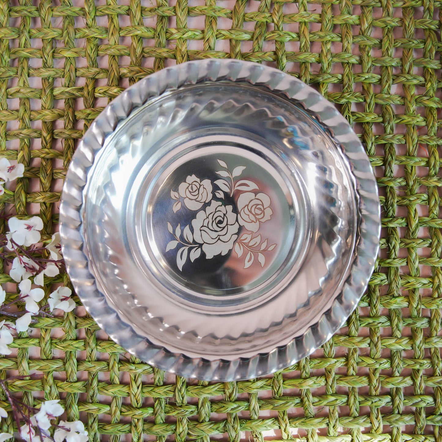 Stainless steel plate with an etched floral design in the center and a contoured rim, placed on a green woven background with white floral accents.