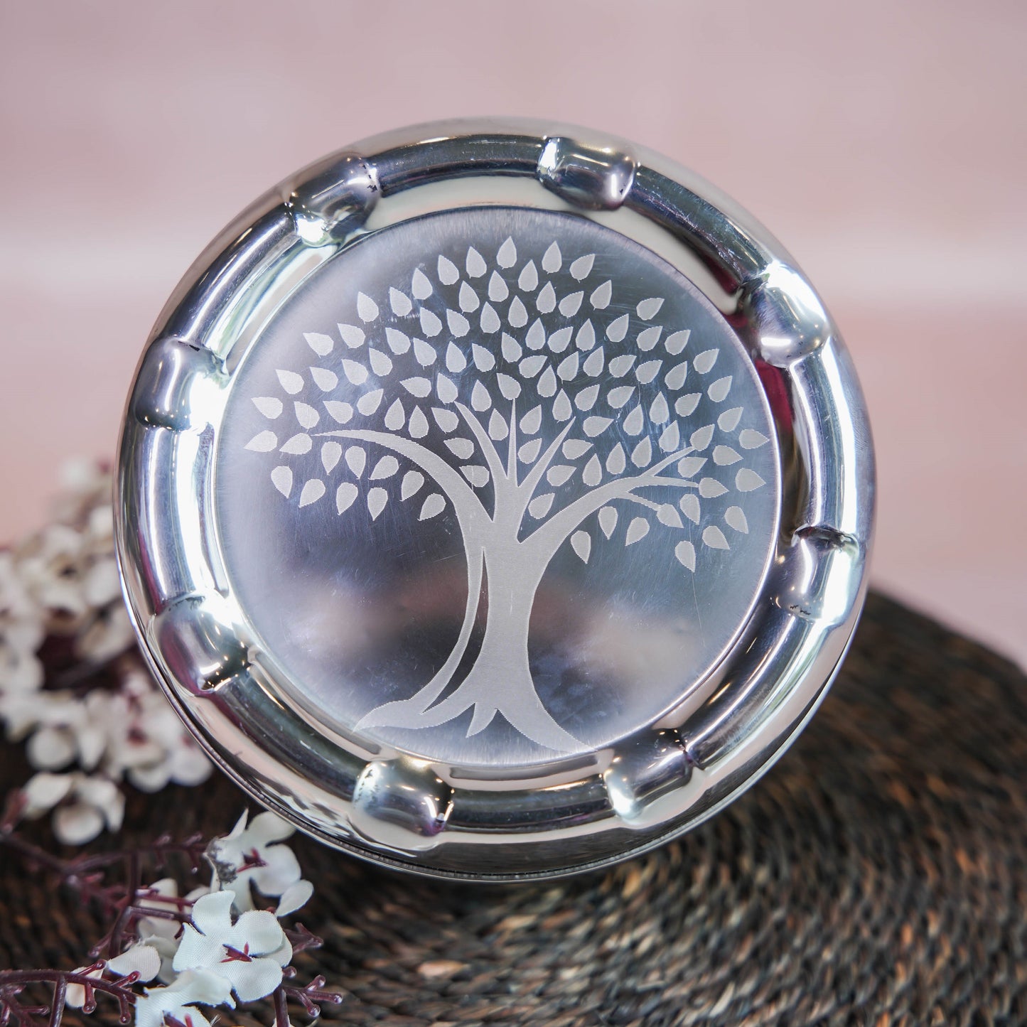 Stainless steel laddu box with a laser-etched tree design on the lid, placed on a woven mat surrounded by floral decor.