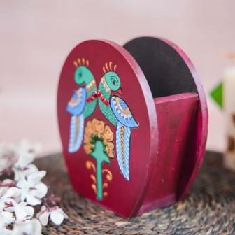 Hand-painted wooden penstand with a design of two colorful parrots perched above a floral motif, placed on a woven mat with decorative floral accents.