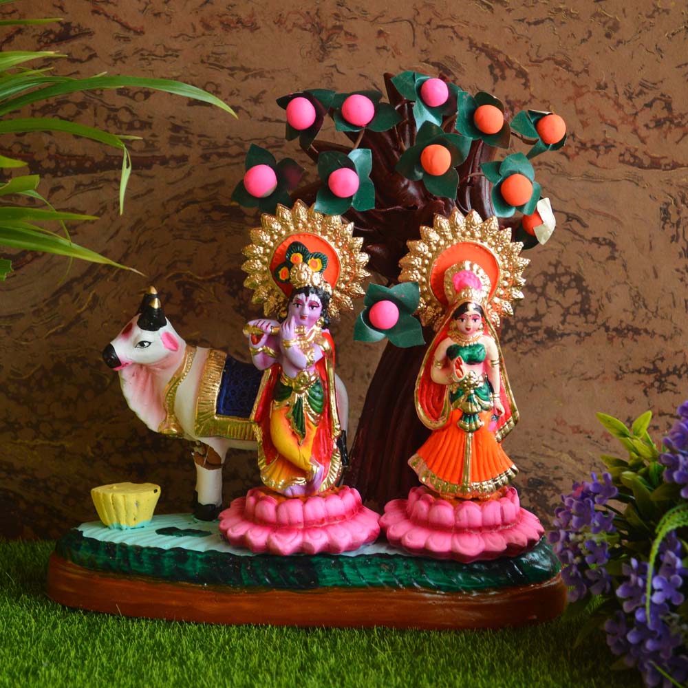 Handcrafted clay dolls of Radha and Krishna standing under a fruit-laden tree, accompanied by a cow, with vibrant colors and intricate detailing.