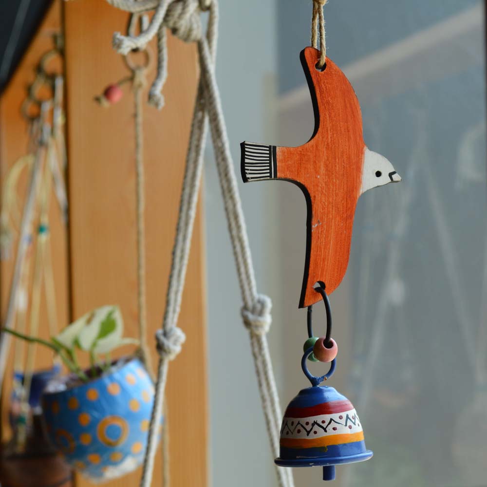 Handmade and hand-painted flying orange bird wind chime with a colorful metal bell, hanging with rustic jute rope and wooden beads near a window with plants.