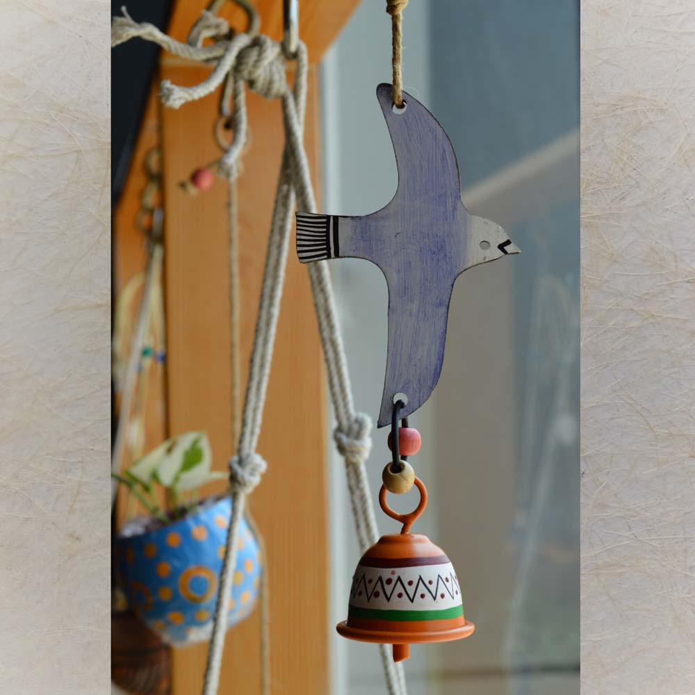 Hand-painted flying blue bird wind chime with a colorful metal bell, hanging on rustic jute rope with wooden beads near a window and plants.