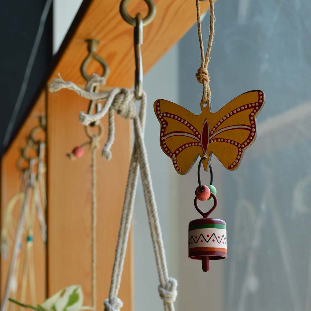 Hand-painted yellow butterfly wind chime with a colorful metal bell, hanging on rustic jute rope with wooden beads, near a sunny window and greenery.
