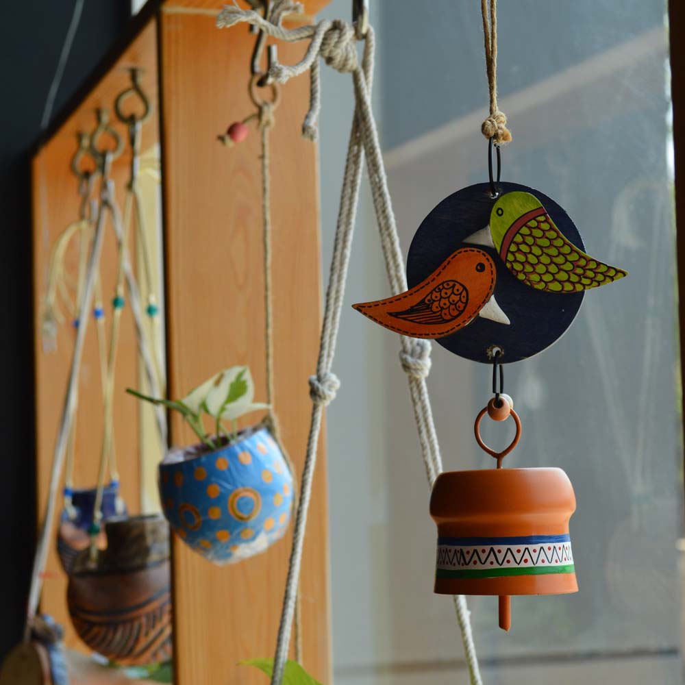 Hand-painted wooden love birds perched on a navy blue circular base with an orange metal bell wind chime, hanging with rustic jute rope and wooden beads near a window with plants.