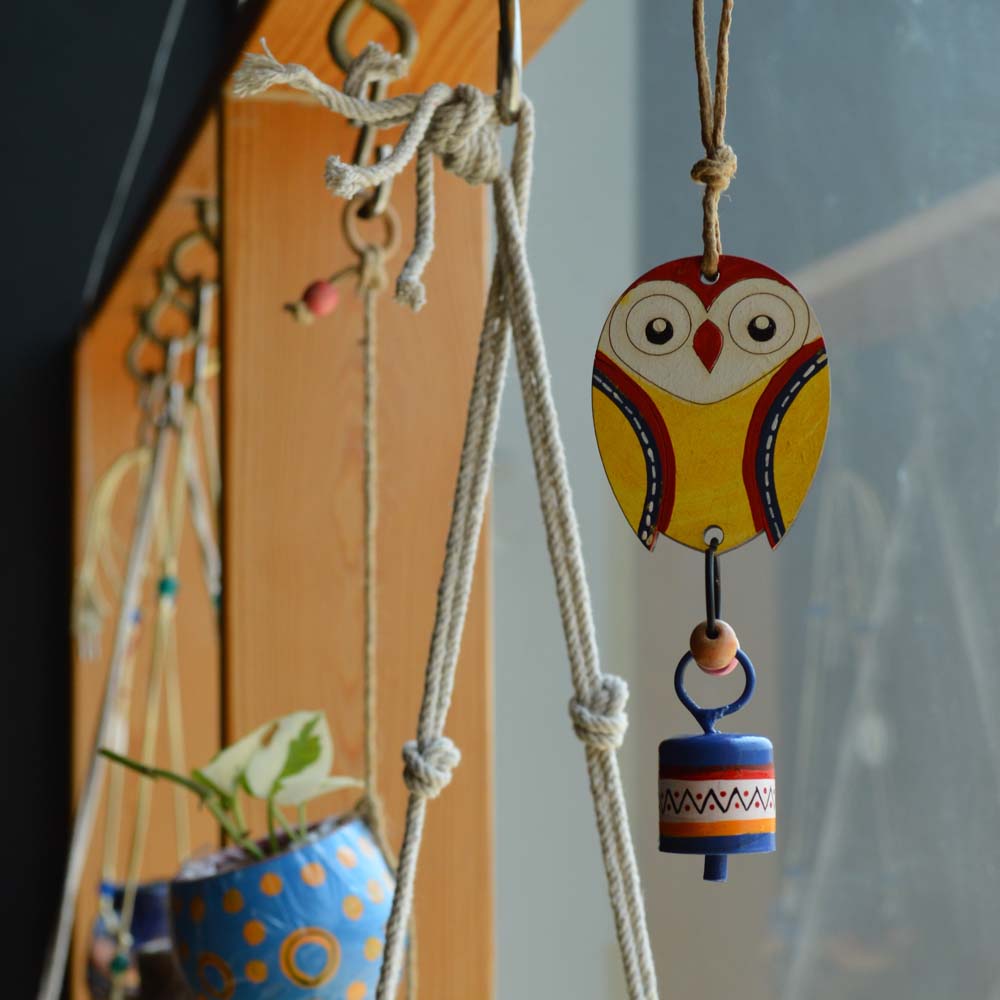 Hand-painted wooden owl in bright yellow with a colorful metal bell wind chime, hanging on rustic jute rope with wooden beads near a window and plants.