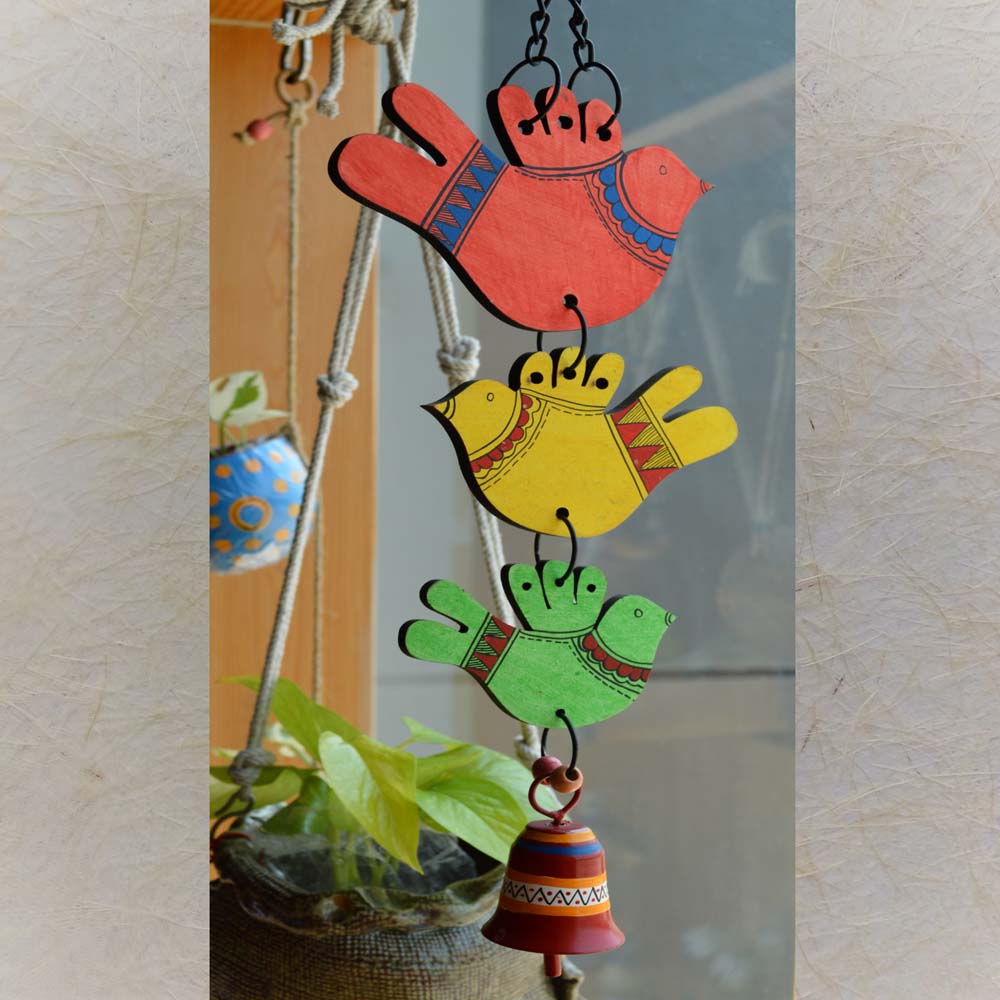 Hand-painted wooden birds in red, yellow, and green with a colorful metal bell wind chime, hanging near a window with plants in the background.