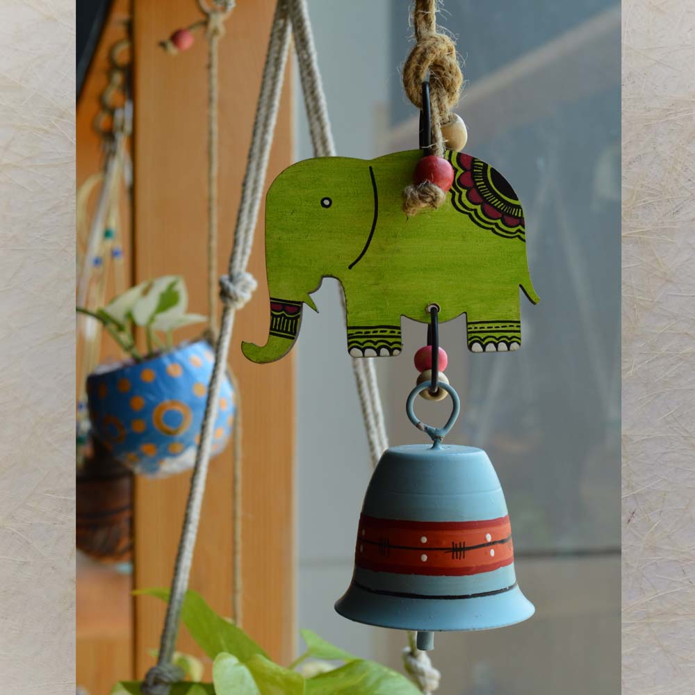 Wooden and metal hand-painted green elephant wind chime with a bell, adorned with jute rope and wooden beads, hanging near a window with plants.
