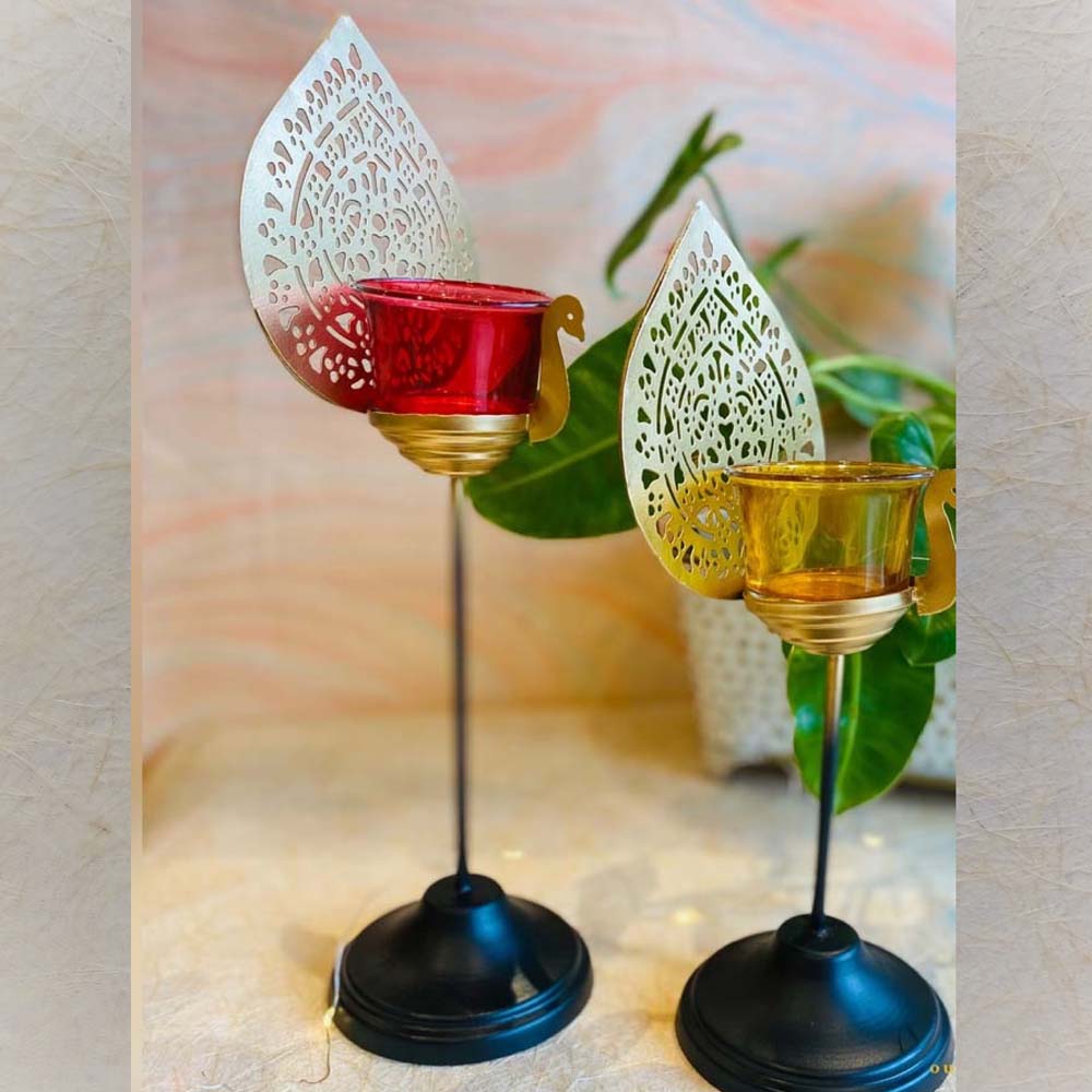 Set of two Peacock Design T-Light Holders with gold-toned intricate feather motifs, vibrant glass holders, and black metal stands, displayed on a wooden surface.