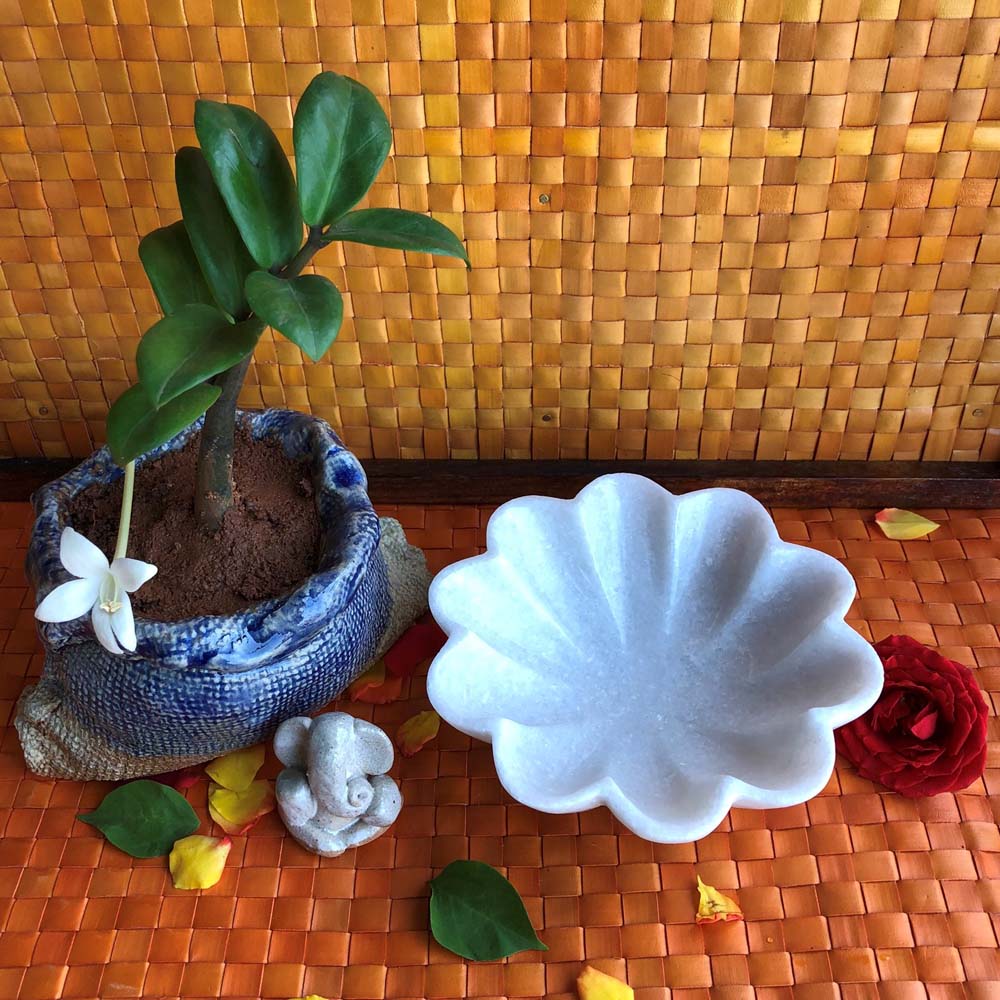 5-inch flower-shaped white marble decorative urli placed next to a potted plant and a small Ganesha idol on an orange woven mat.