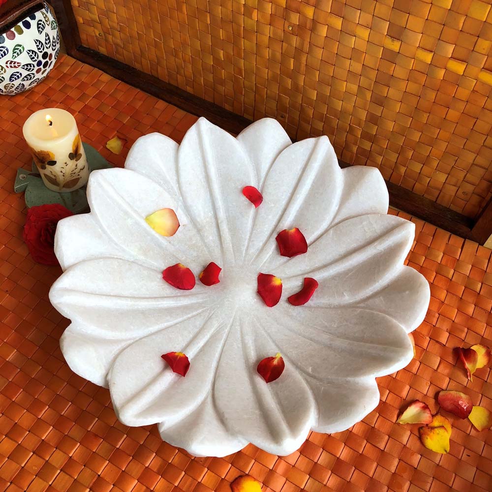 Large white marble lotus flower-shaped decorative urli with scattered red rose petals, placed on an orange woven mat with a candle in the background.