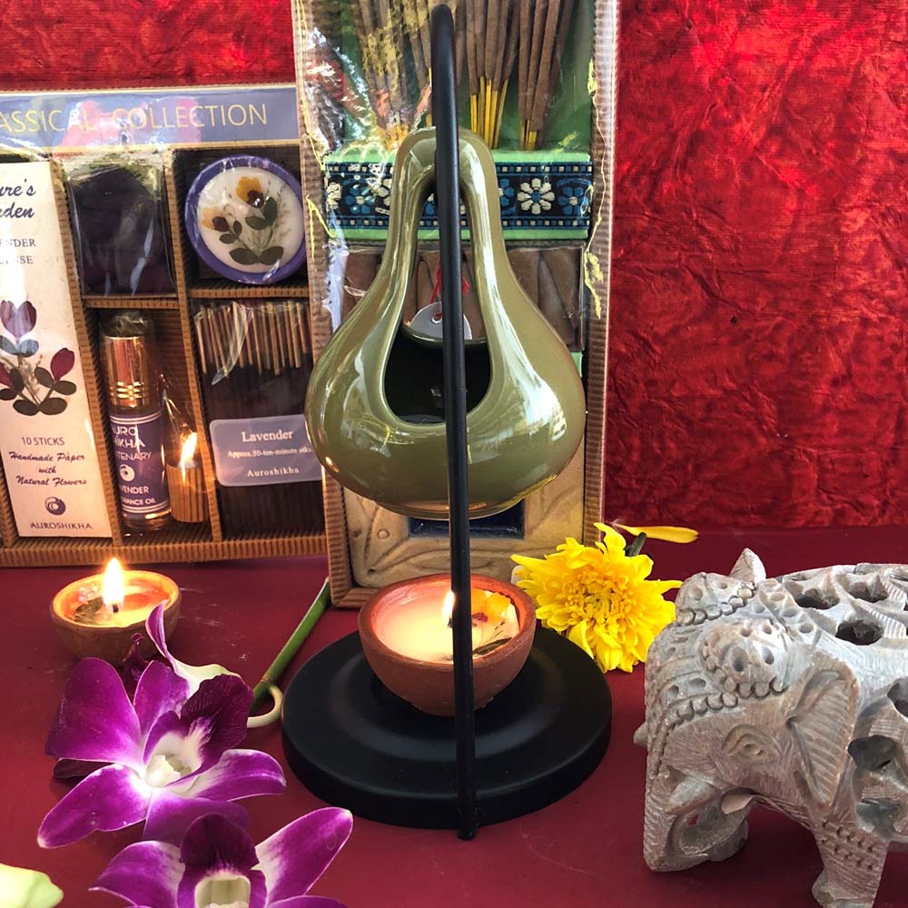 Cera Green Ceramic Oil Diffuser with a lit tealight, surrounded by incense sticks, flowers, and a carved elephant figurine on a vibrant red background.