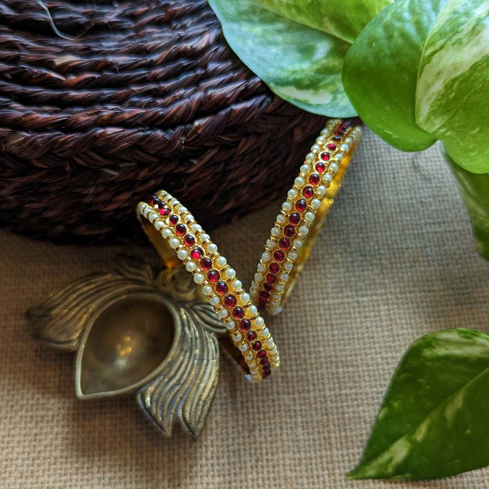 Gold bangles with ruby red stones and pearl detailing, placed on a rustic background with green leaves and brass decor.