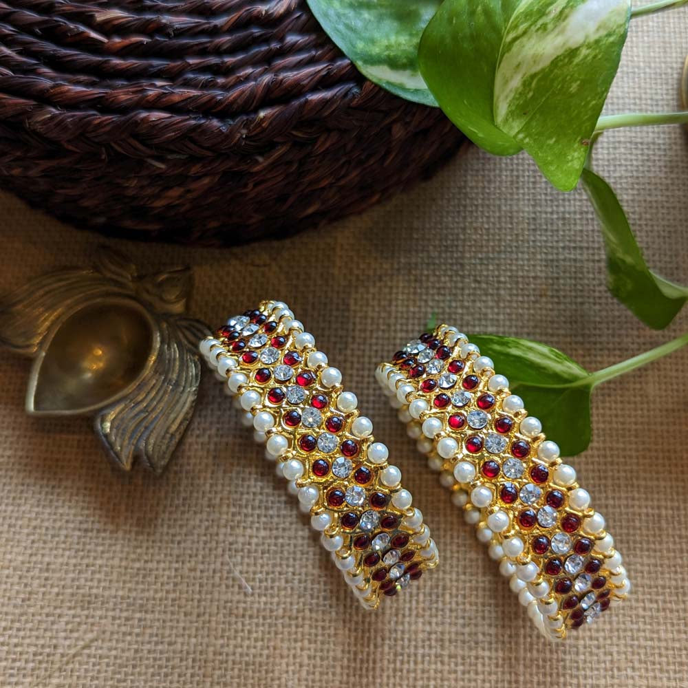 Gold bangles with ruby red stones and pearl detailing, placed on a rustic background with green leaves and brass decor.