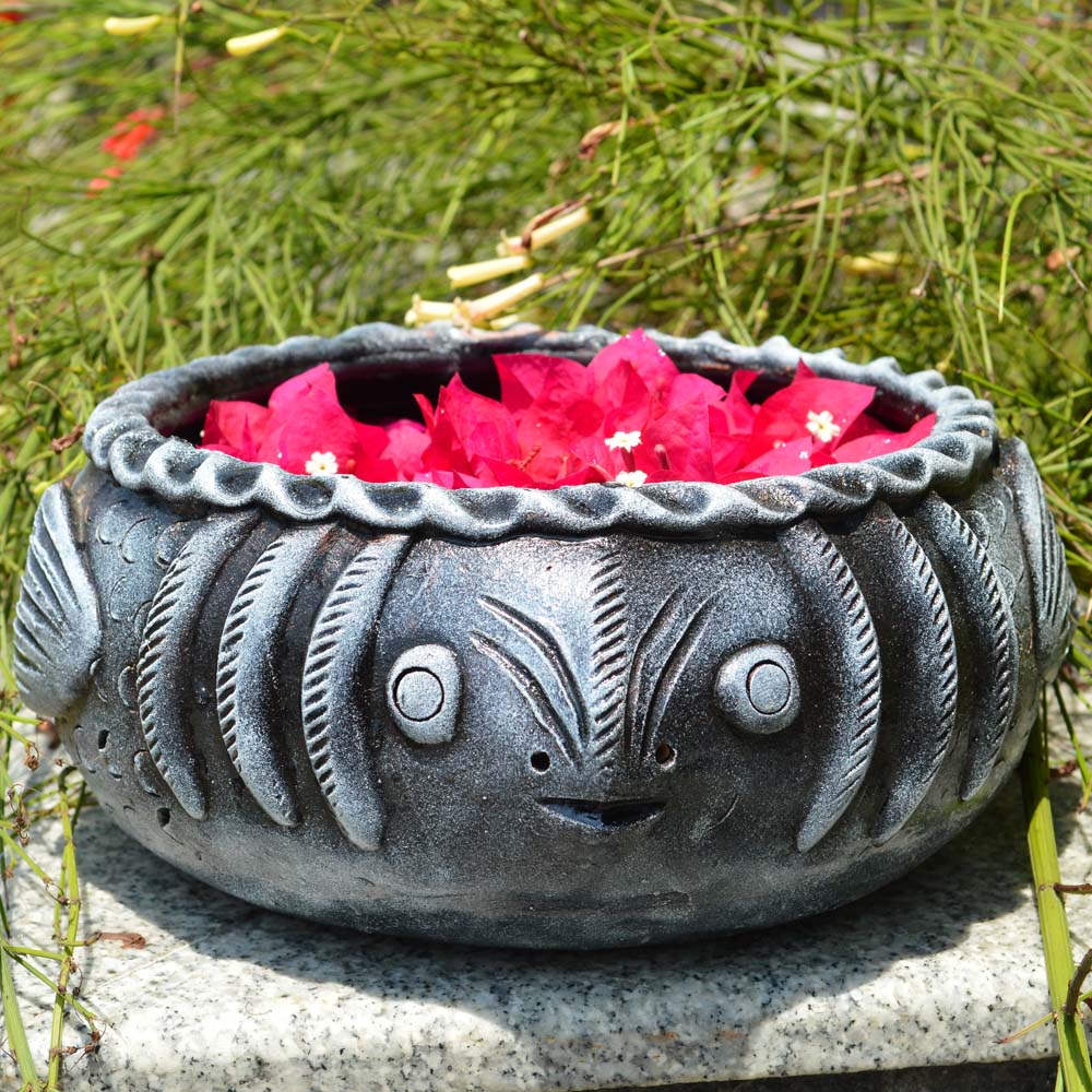 Handcrafted stone-finish fish-shaped earthen urli filled with vibrant pink bougainvillea petals, set against a lush outdoor backdrop.