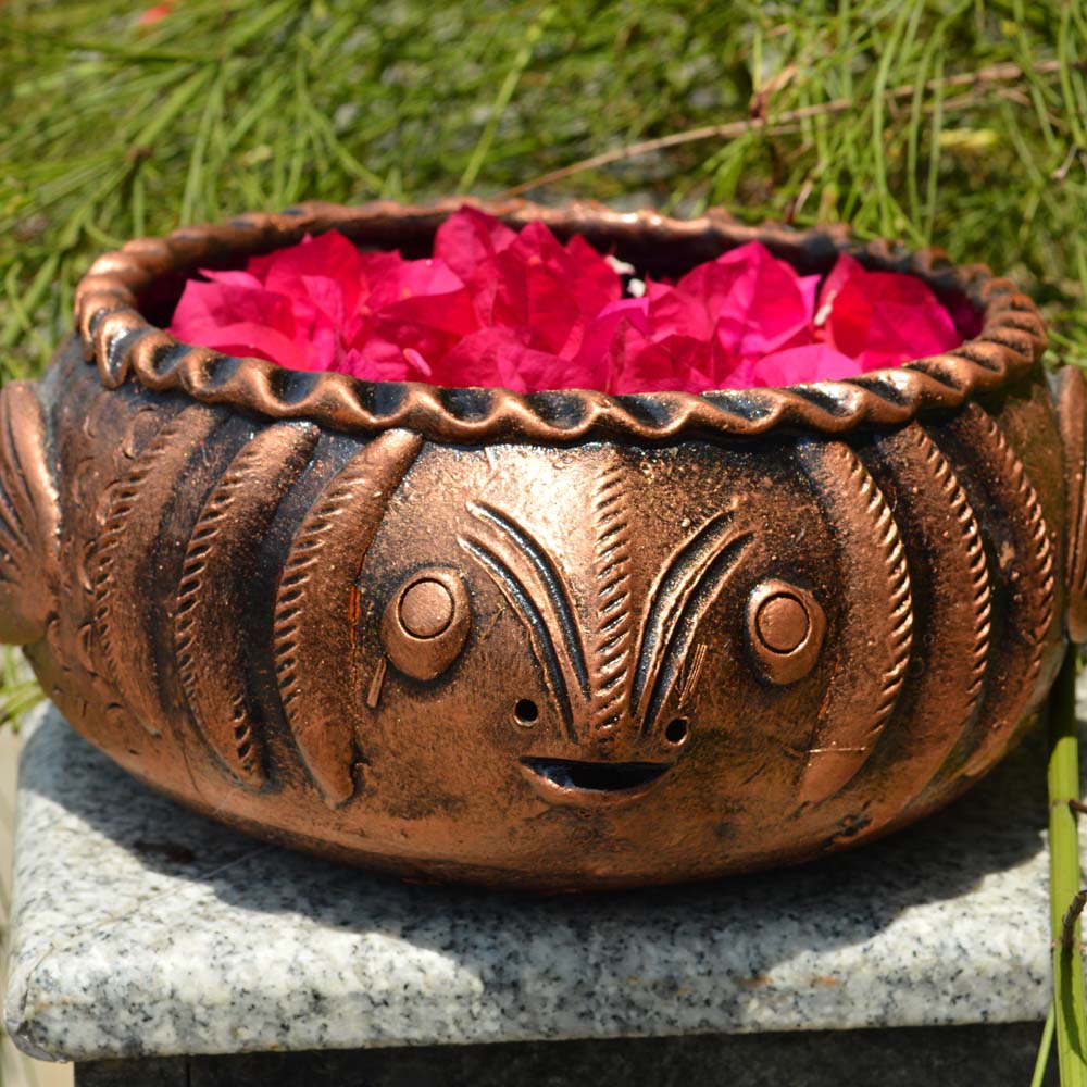 Handcrafted copper-finish fish-shaped earthen urli filled with vibrant pink bougainvillea petals, placed outdoors amidst lush greenery.
