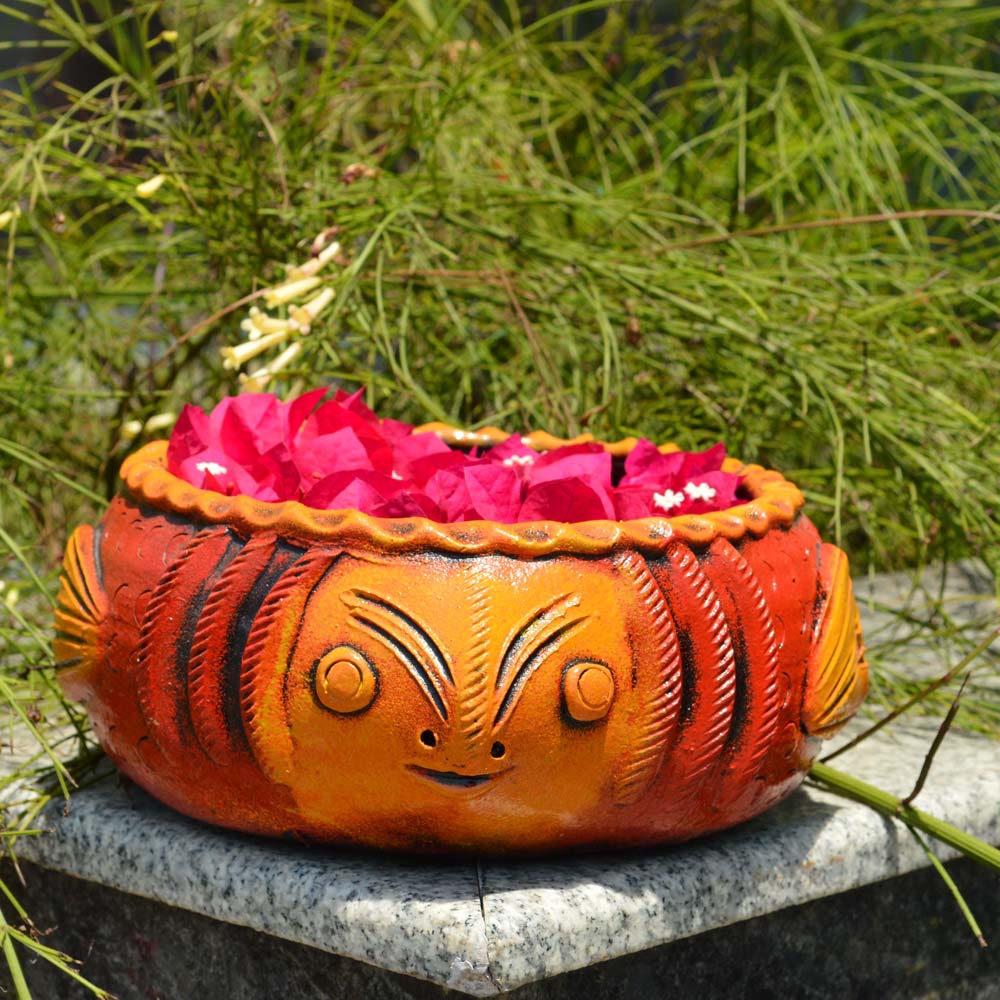 Bright orange handcrafted fish-shaped earthen urli filled with vibrant pink bougainvillea petals, set against a lush green outdoor backdrop.