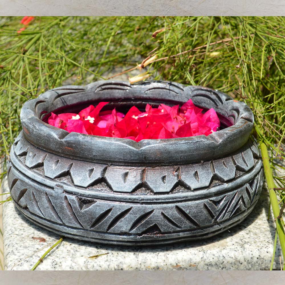 Small earthen urli with a stone-textured finish, filled with vibrant pink bougainvillea petals, displayed in an outdoor garden setting.