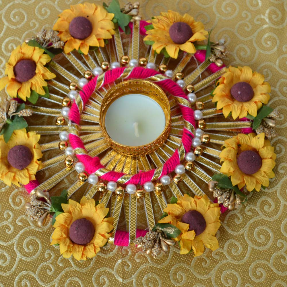 Round sunflower-themed T-light diya with yellow artificial flowers, pearls, and a golden lattice holder on a pink and gold base.