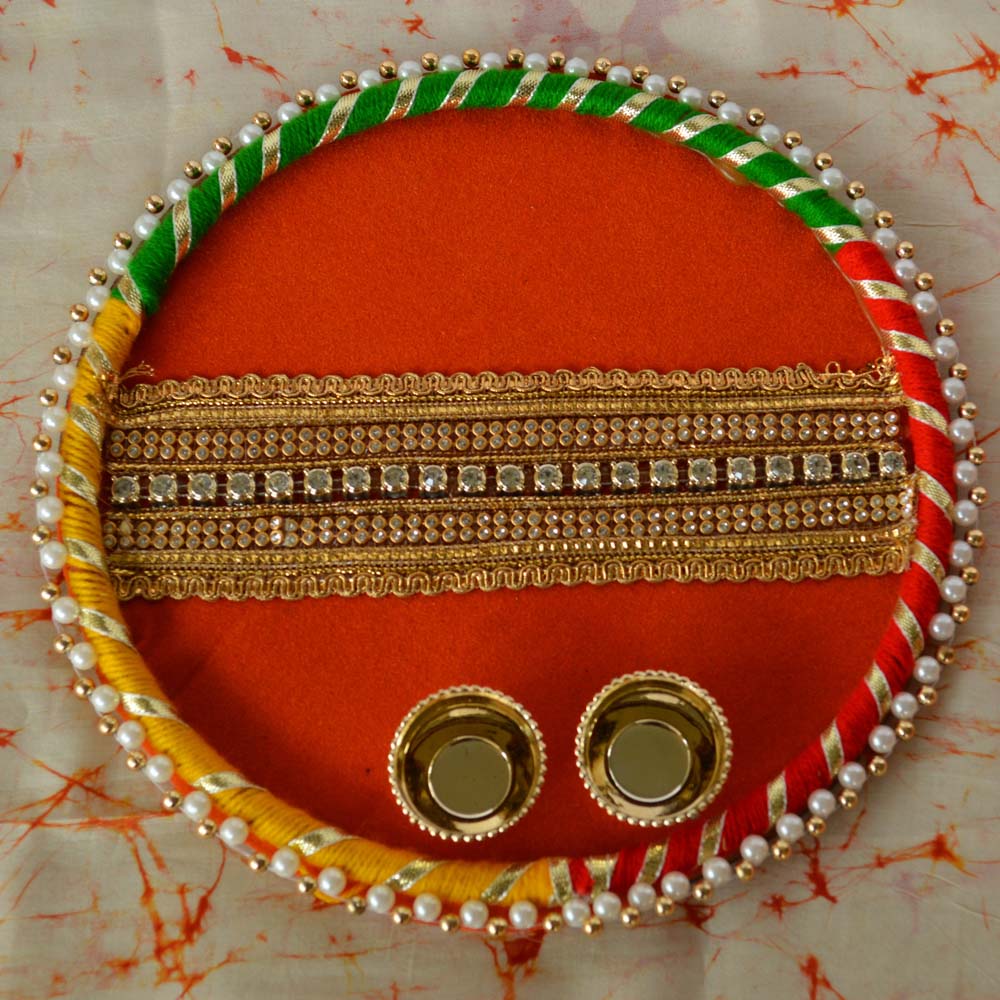 Round kumkum plate with orange base, colorful borders, pearl accents, golden embroidery, and two brass cups, ideal for pooja and festive use.