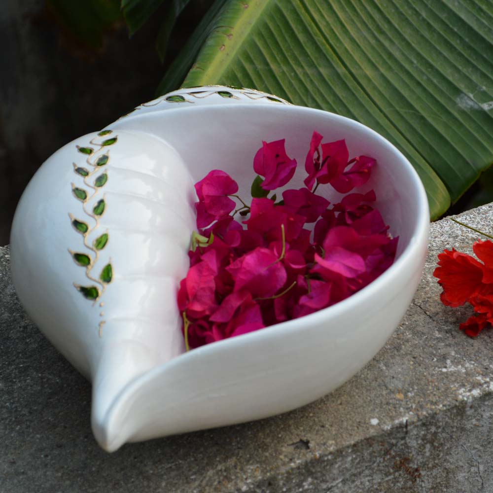 White earthen Sanghu Urli in the shape of a conch shell, filled with vibrant pink bougainvillea petals, placed outdoors.