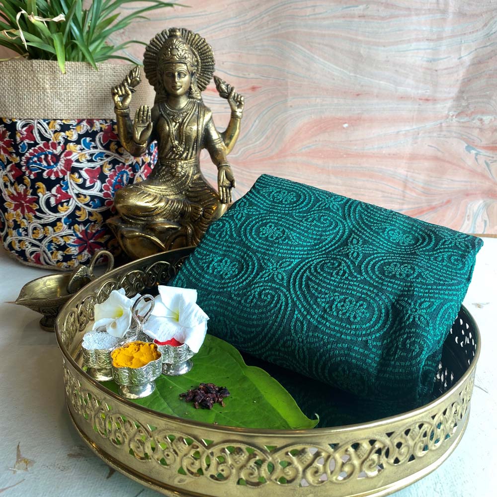 Banaras dark green brocade fabric with intricate golden swirling motifs, styled with traditional brass decor and floral arrangements.