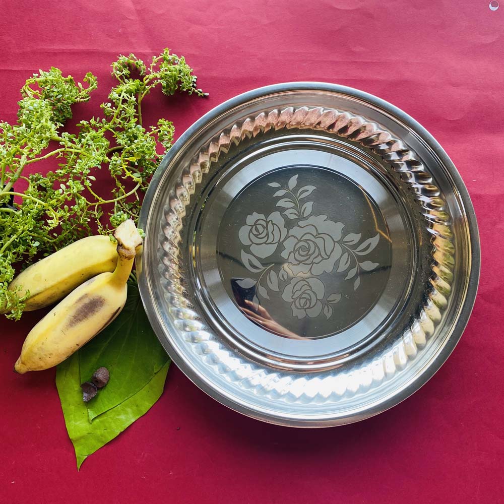 Stainless steel puja plate with floral engraving, placed on a red background, surrounded by green leaves and bananas.