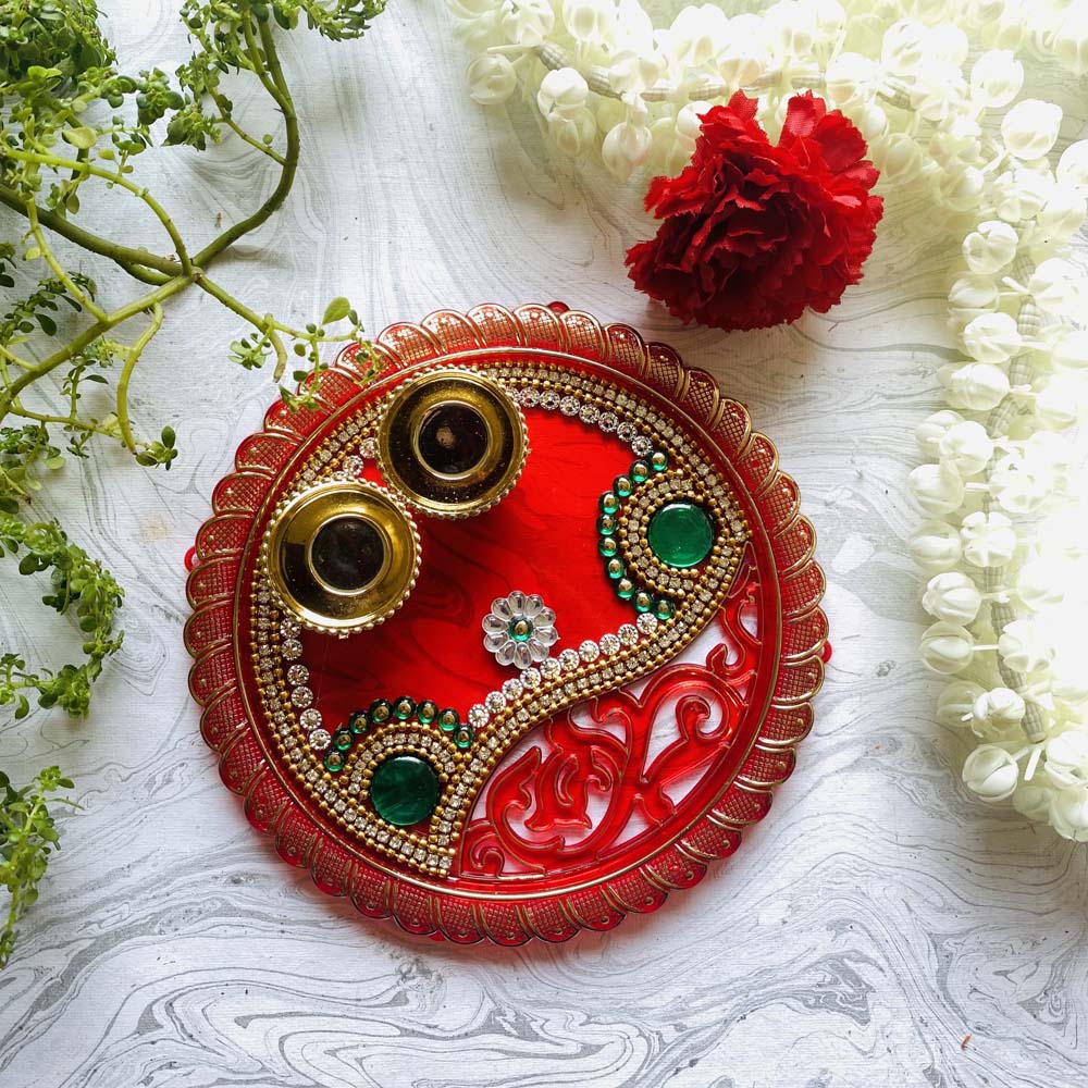 Red decorative thali with two brass cups, adorned with stone embellishments, green accents, and floral detailing, ideal for pooja or festive use.