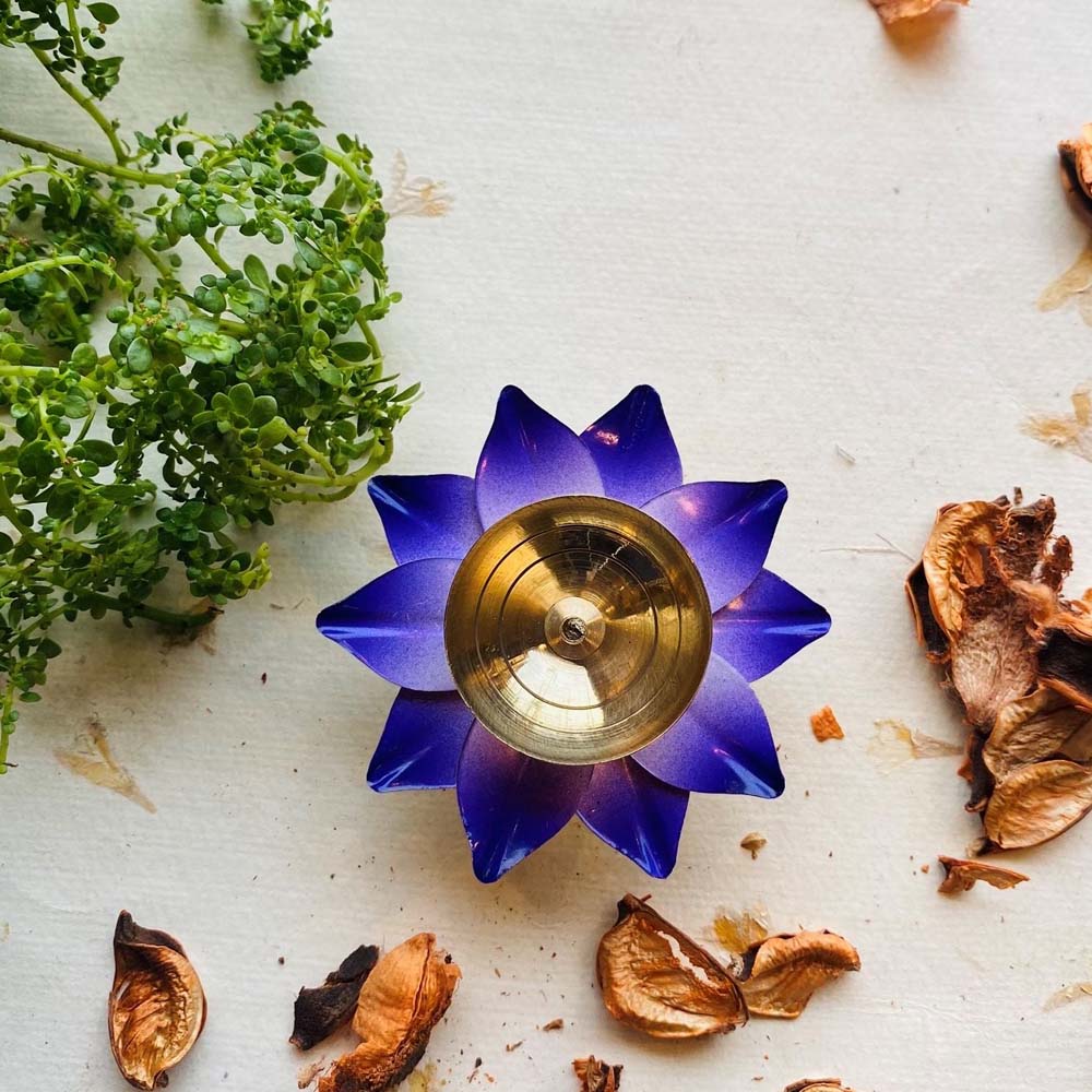 Handcrafted purple lotus diya with a brass center, surrounded by greenery and dried petals on a white textured surface.