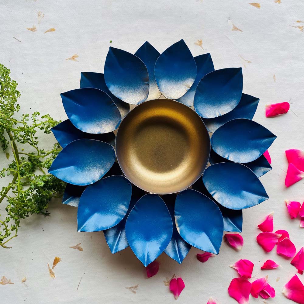Blue decorative urli with a gold hammered metal bowl and bright blue lotus petals, surrounded by scattered pink rose petals, placed on a neutral background with greenery.