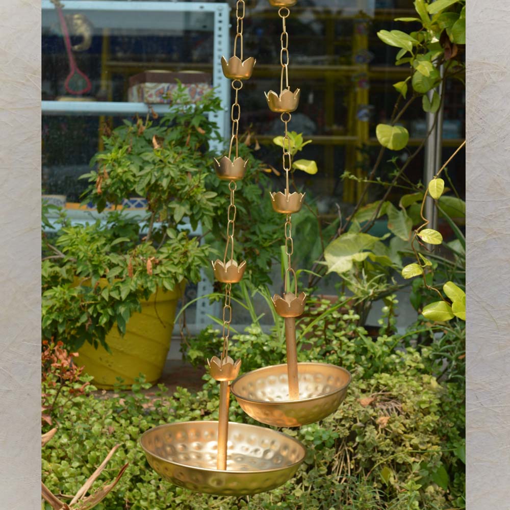 Set of 2 decorative hanging gold urlis with hammered bowls and floral chain design, displayed outdoors in a lush green setting.
