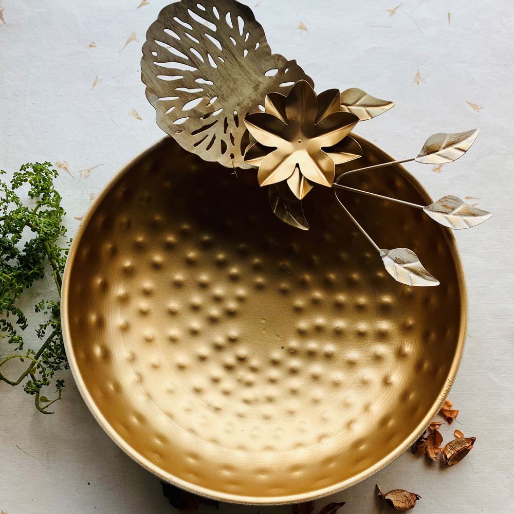 Large gold decorative urli with a hammered metal bowl and intricate floral and leaf accents, placed on a neutral background with greenery and potpourri.
