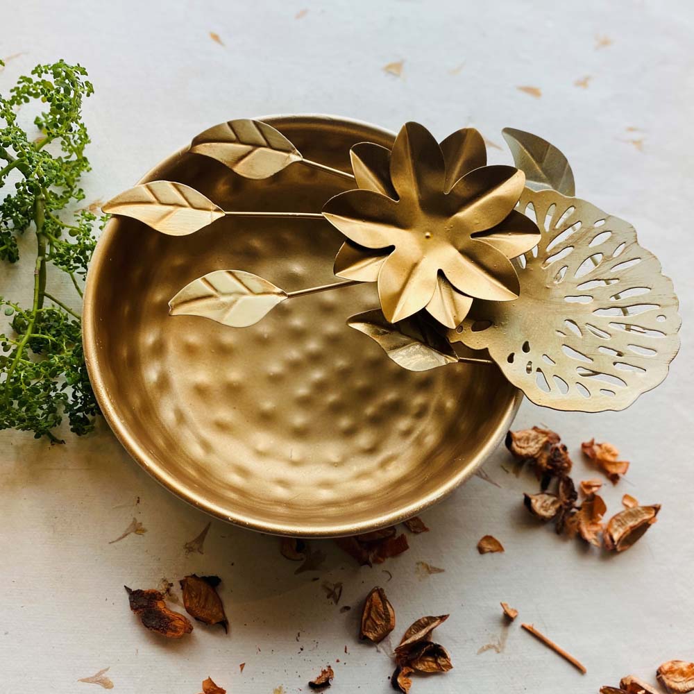 Medium-sized gold decorative urli with a hammered metal bowl, featuring floral and leaf designs, placed on a neutral background with greenery and potpourri.