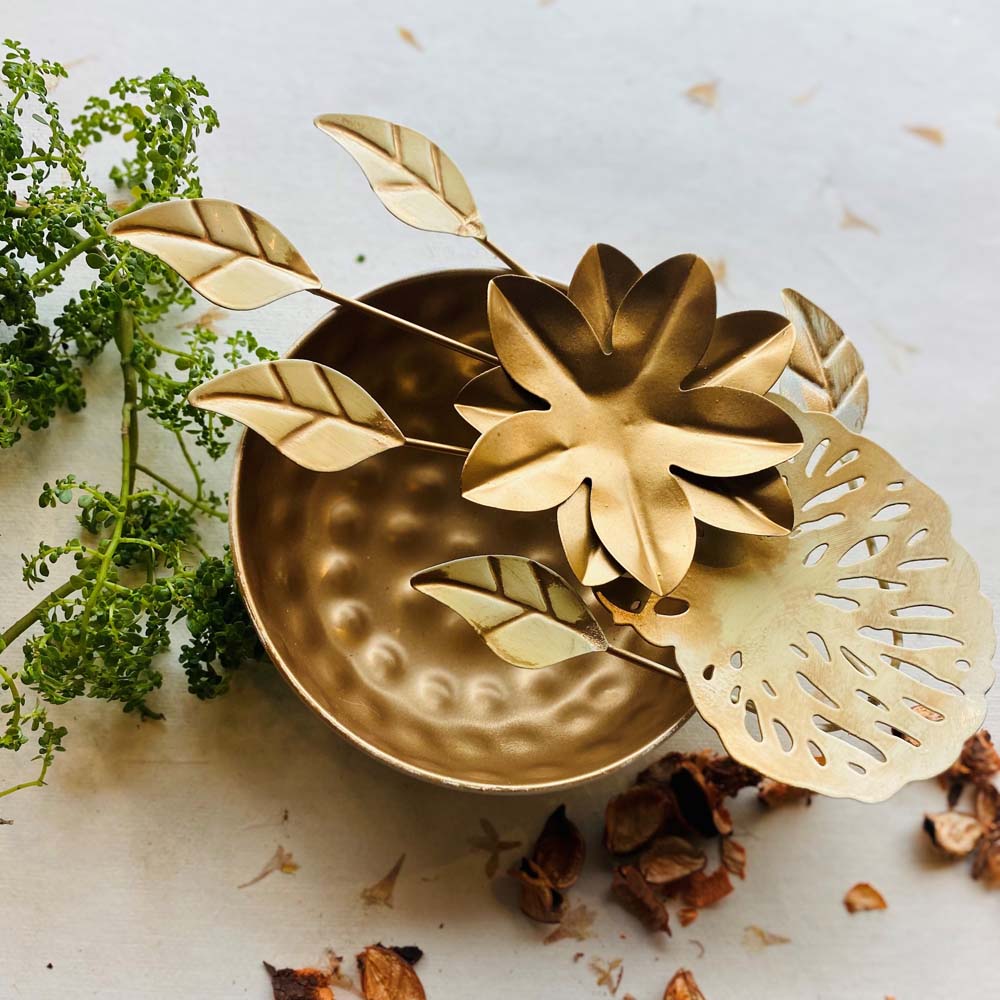 Small gold decorative urli with a hammered metal bowl, adorned with floral and leaf designs, placed on a neutral background with natural greenery and potpourri.