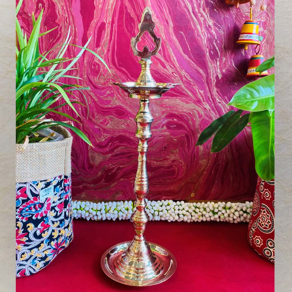 Traditional brass kuthu vilakku (oil lamp) with an intricate top design, surrounded by plants and vibrant decor on a red surface.