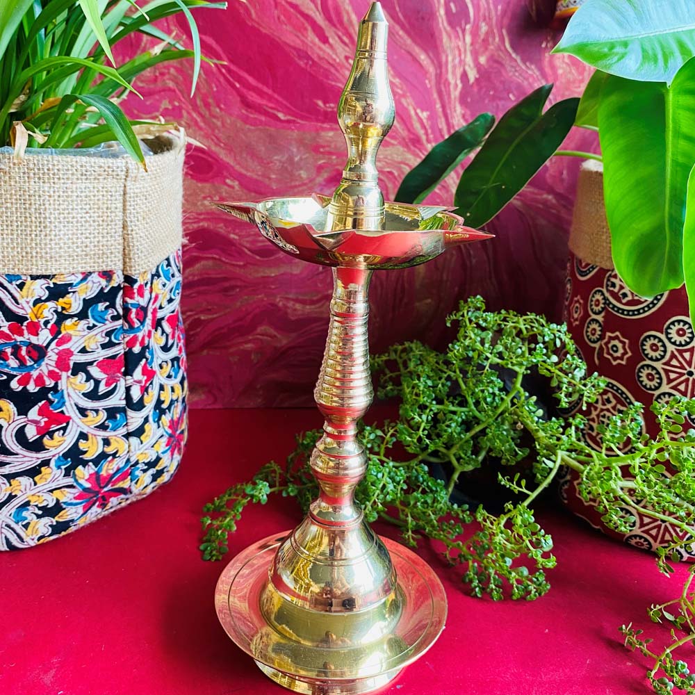 Handcrafted brass Kerala lamp (Nilavilakku) with a polished finish, placed on a vibrant red surface with plants and colorful decor in the background.