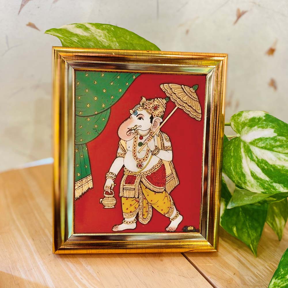 Tanjore-style artwork of Lord Ganesha holding an umbrella and lamp, set against a red and green backdrop in a gold-colored frame.