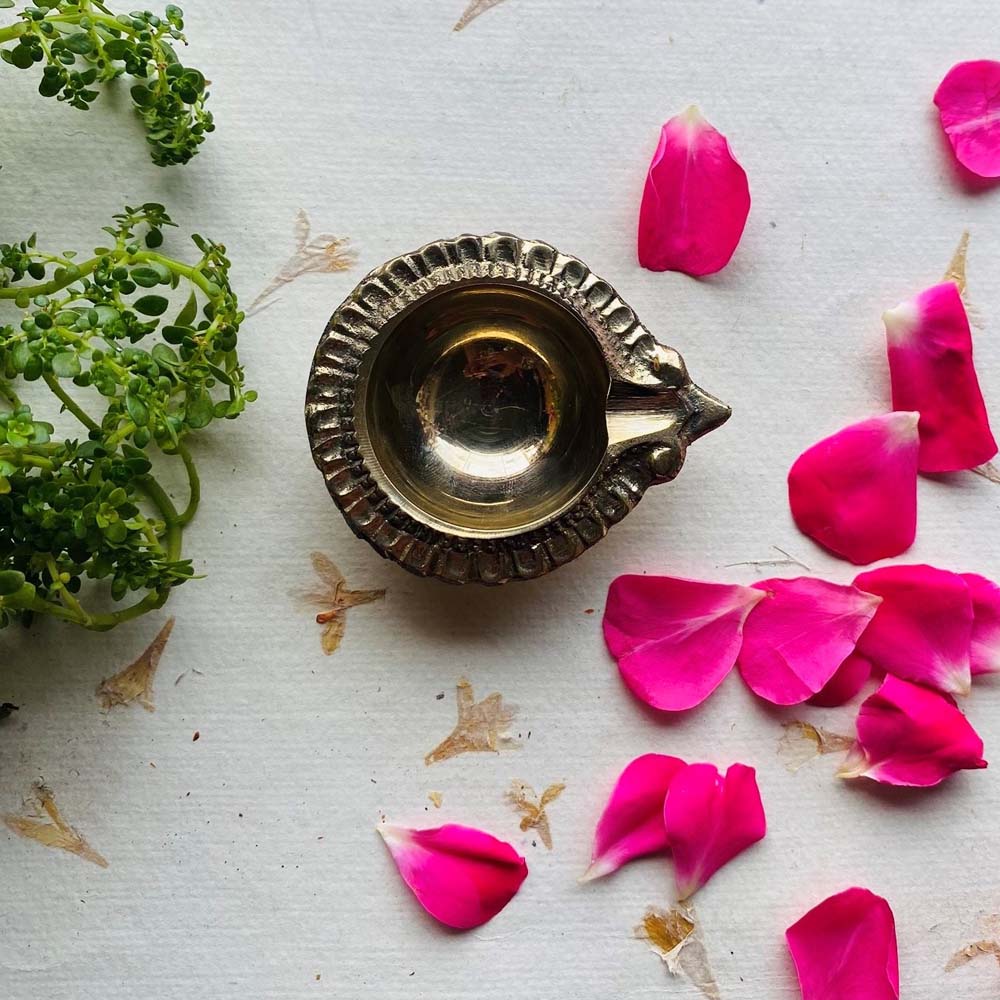 A beautifully crafted brass diya (oil lamp) surrounded by pink rose petals and greenery on a white textured surface.