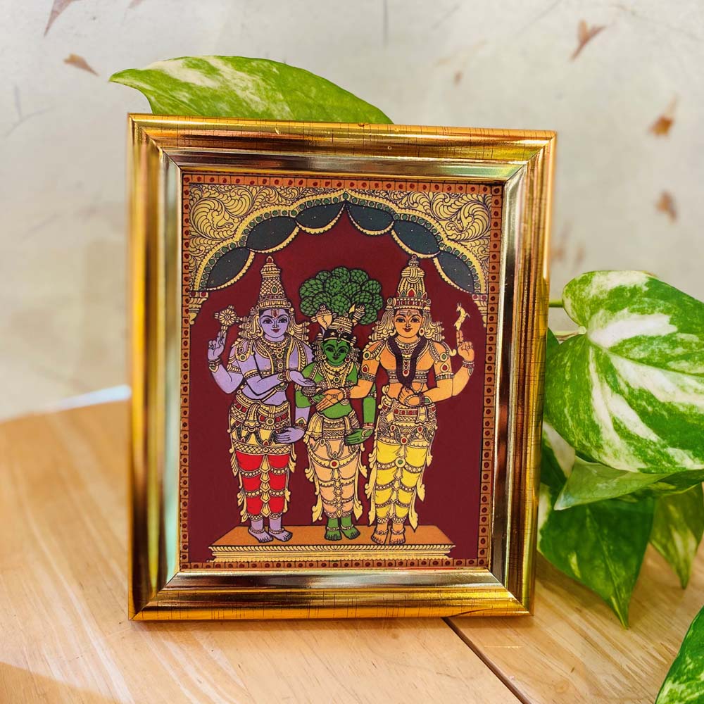 Tanjore-style artwork of Meenakshi Kalyanam featuring Lord Sundareswarar, Goddess Meenakshi, and Lord Vishnu, set against a maroon backdrop in a gold frame.