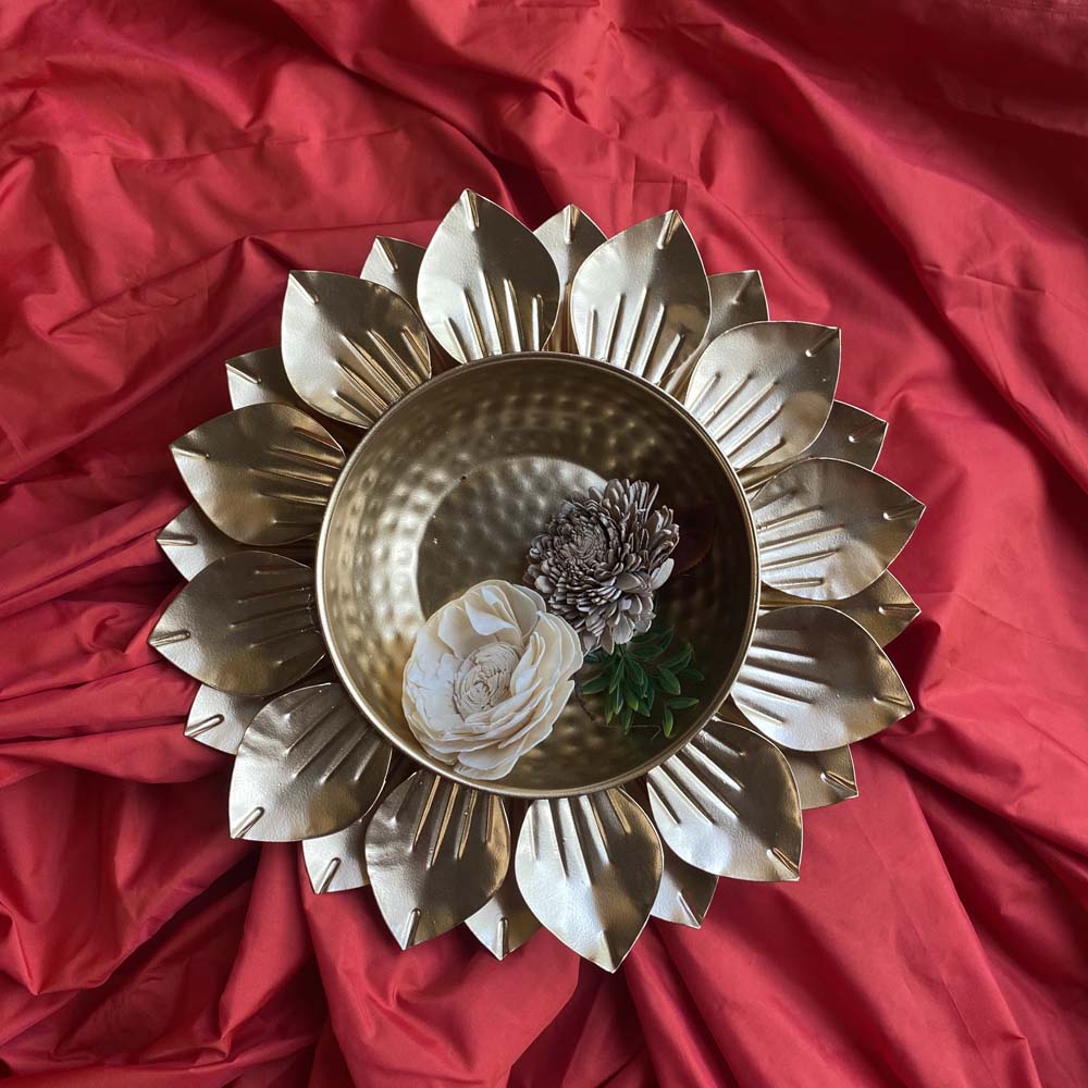 Small gold lotus-shaped decorative urli with a hammered metal bowl, adorned with artificial flowers, placed on a vibrant red fabric.
