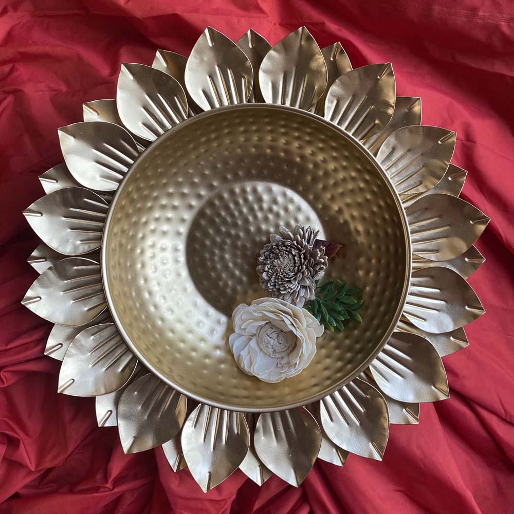 Gold lotus-shaped decorative urli with a hammered metal bowl, adorned with floral arrangements, placed on a vibrant red backdrop.