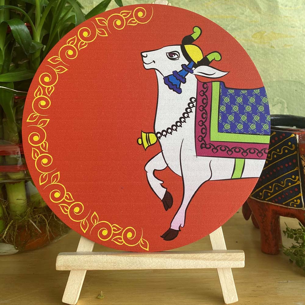 Hand-painted decorative plate with a white cow adorned in traditional patterns, set against an orange background with a golden ornamental border, displayed on a wooden easel.