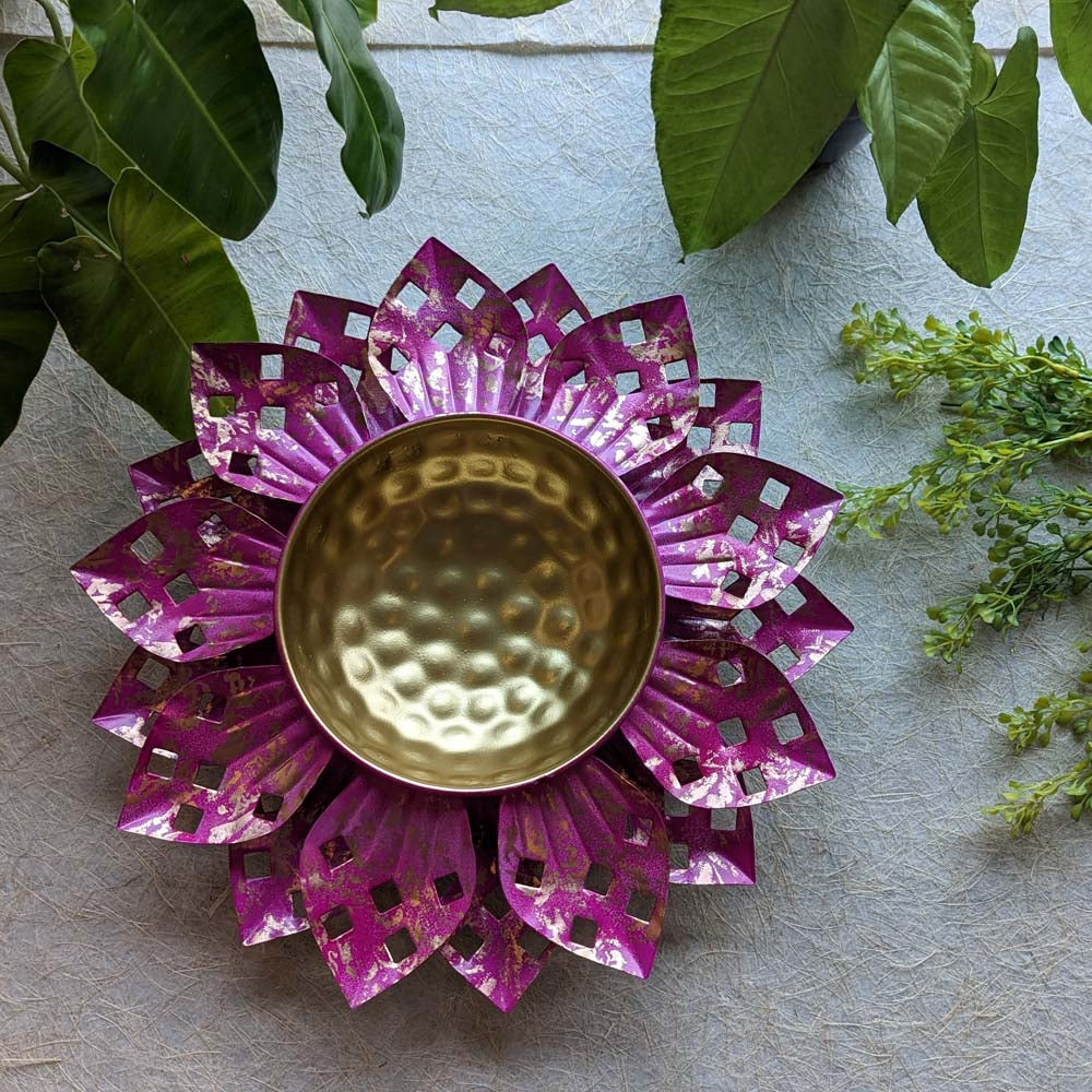 Handcrafted floral metal urli with pink petals and a golden hammered center bowl, placed among green plants.
