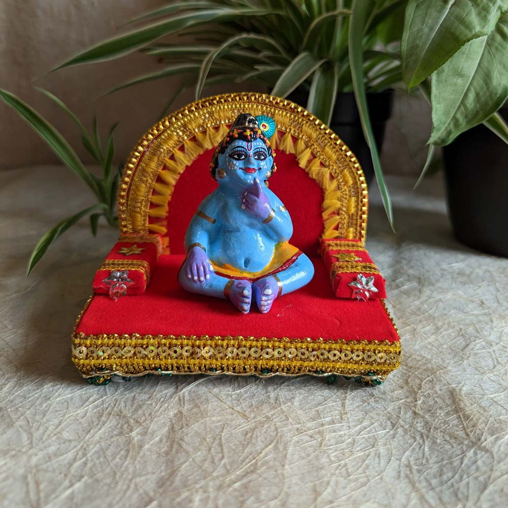 Handcrafted Laddu Krishna idol seated on a decorated Simhasan with red and gold embellishments.