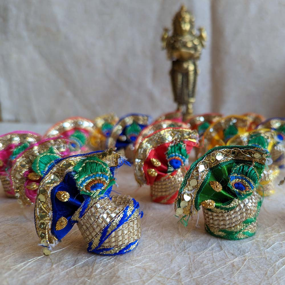 Vibrant handcrafted decorative turbans with embroidered peacock feather motifs, zari detailing, and gold accents, displayed with a brass deity statue.