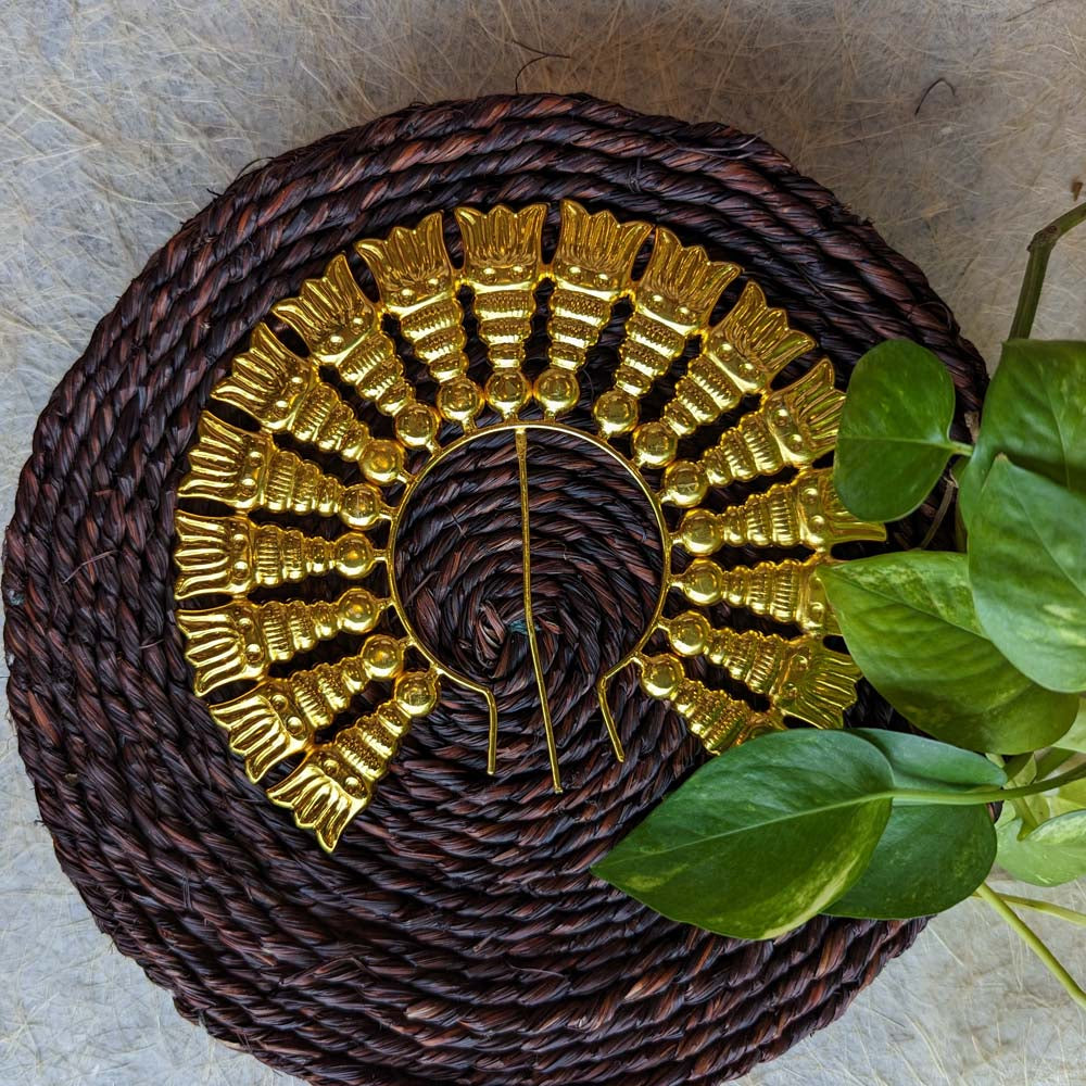 Gold-toned Jada Billai with intricate circular design placed on a brown woven mat with green leaves.